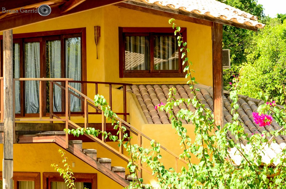 Fachada do chalé com paredes amarelo-ocre, madeira escura e vegetação tropical com buganvília